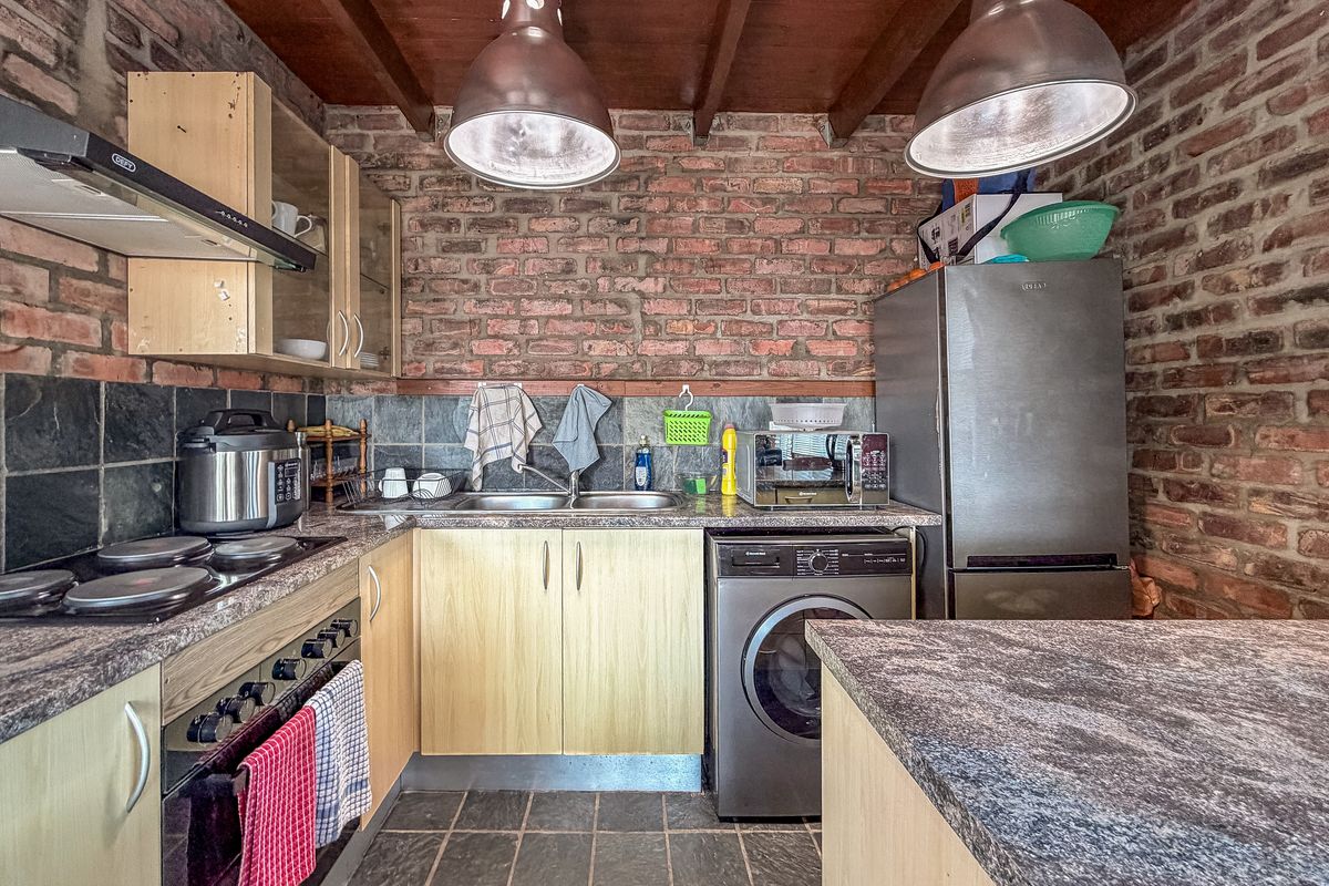 A rustic kitchen featuring exposed brick walls, slate tile flooring, timber ceiling beams, and functional cabinetry with integrated appliances.