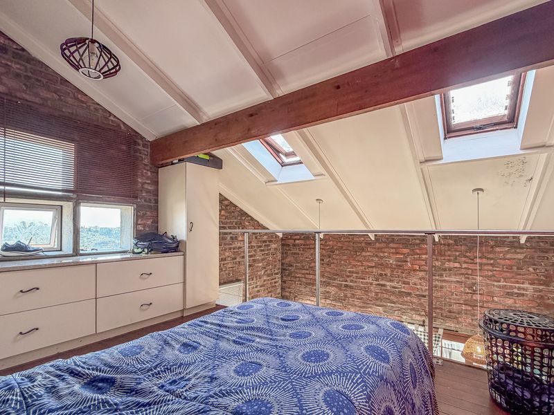 A loft-style bedroom featuring exposed brick walls, timber ceiling beams, skylights, and a modern metal balustrade overlooking the lower level.