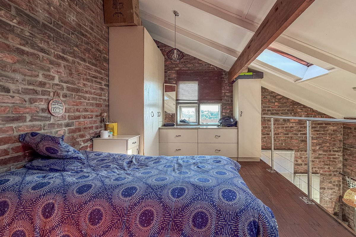 A characterful loft-style bedroom featuring exposed brick walls, timber ceiling beams, a skylight, and built-in storage, overlooking the lower level via a metal balustrade.