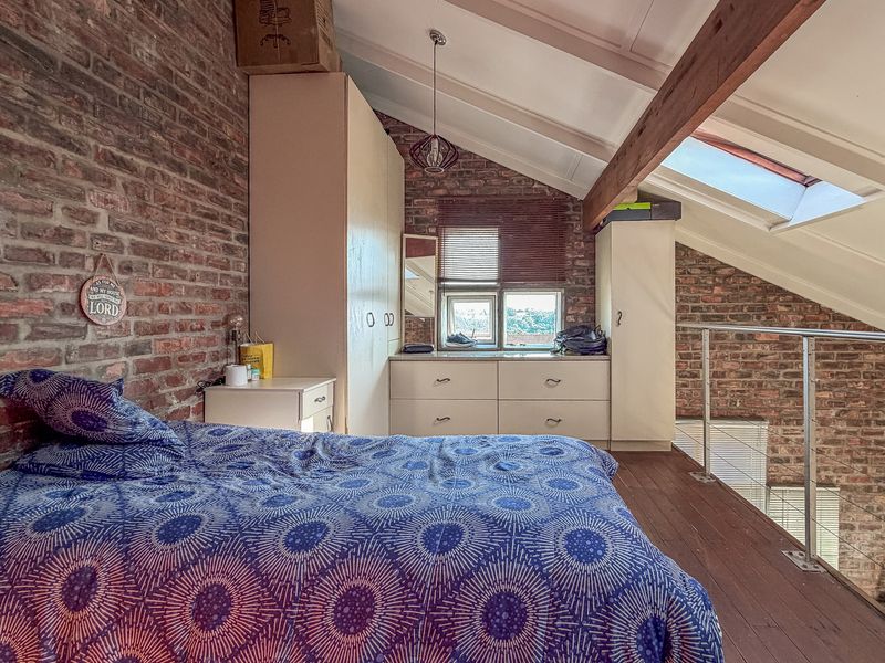 A characterful loft-style bedroom featuring exposed brick walls, timber ceiling beams, a skylight, and built-in storage, overlooking the lower level via a metal balustrade.