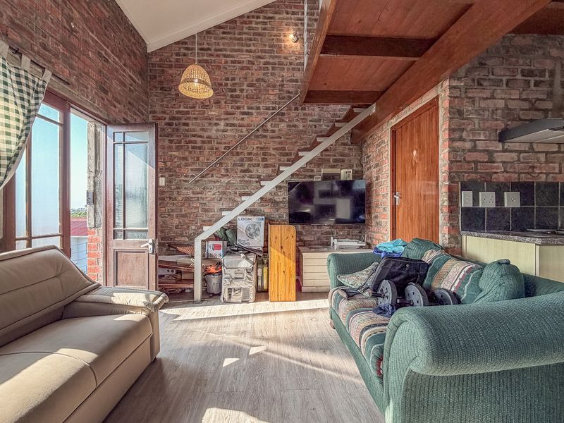 Open-plan living area featuring exposed brick walls, a timber staircase, vaulted ceilings, and an integrated kitchen space with tiled backsplash.