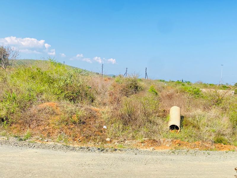 A view of an undeveloped residential plot featuring natural vegetation, utility poles, and a drainage pipe, accessible from a gravel road.