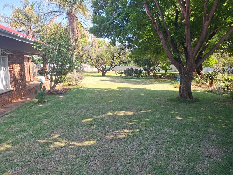 A spacious, well-maintained garden featuring mature trees, a lush lawn, and a view of the home's brick exterior and covered patio area.