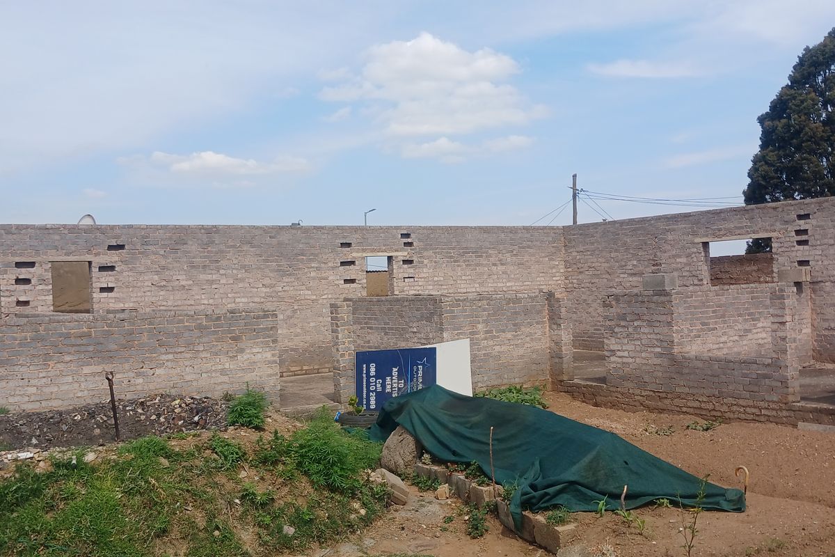 An unfinished residential construction site showing exposed brick masonry walls and open window frames in a rear yard setting.