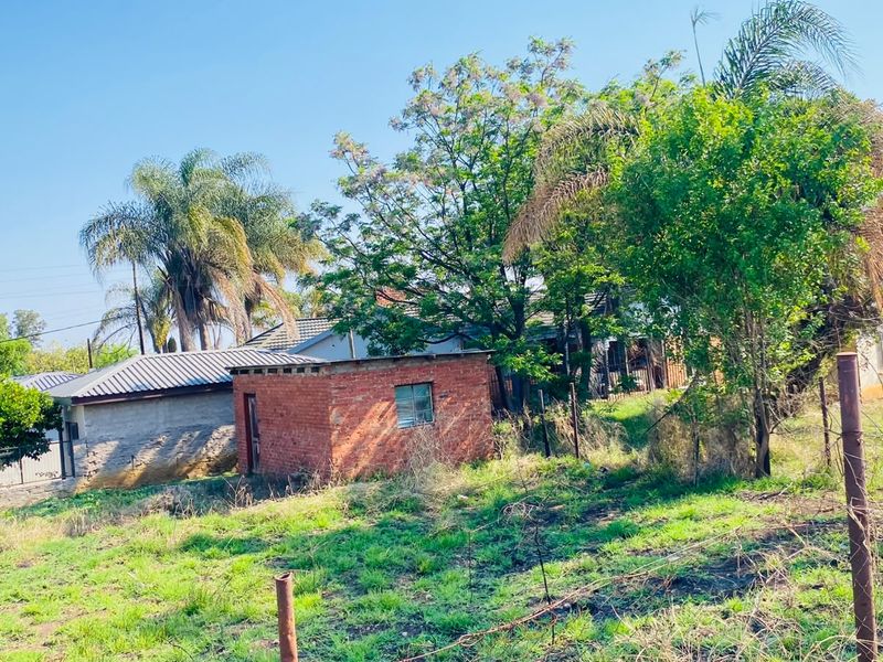 A view of a residential plot featuring a small brick outbuilding, mature trees, and perimeter wire fencing on a grassy lot.