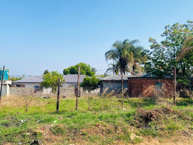 A vacant residential plot featuring wire fencing, metal posts, and a water storage tank, with surrounding neighborhood structures visible in the background.