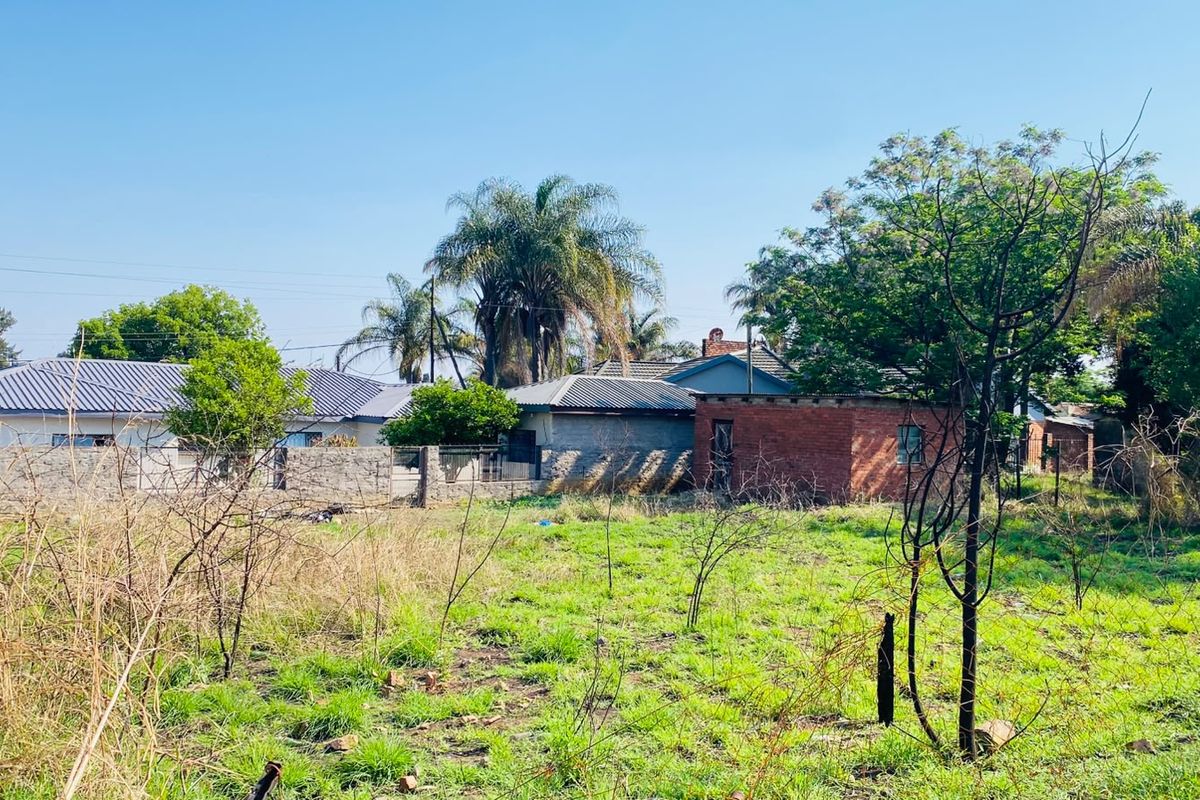 A view of a vacant residential plot featuring overgrown grass, wire fencing, and a brick boundary wall with neighboring properties in the background.
