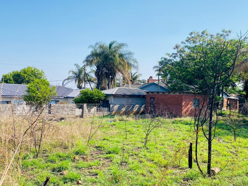 A view of a vacant residential plot featuring overgrown grass, wire fencing, and a brick boundary wall with neighboring properties in the background.