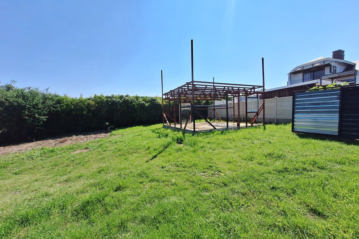 A residential garden area featuring a grassy lawn, perimeter hedging, and an unfinished metal structural frame for a potential outdoor building or gazebo.