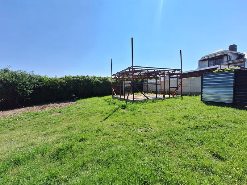 A residential garden area featuring a grassy lawn, perimeter hedging, and an unfinished metal structural frame for a potential outdoor building or gazebo.