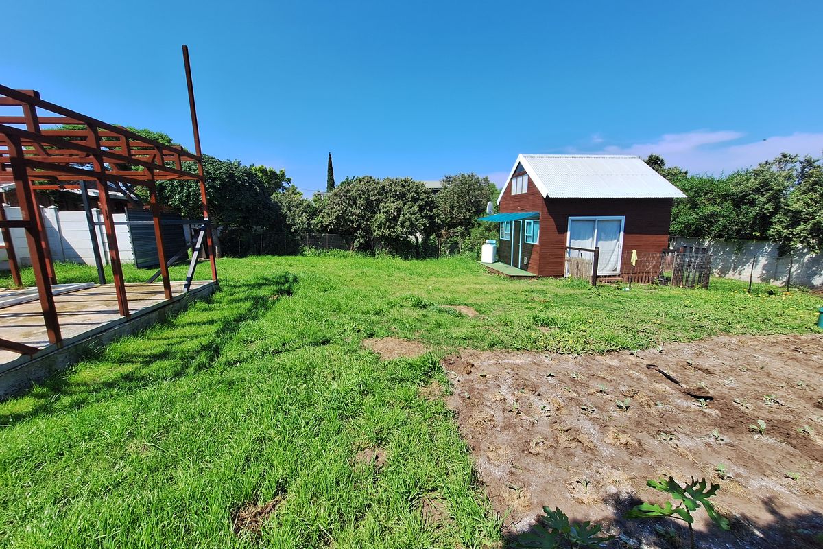 A spacious residential garden featuring a small wooden cabin, an unfinished timber frame structure, and a mix of lawn and cultivated soil areas.