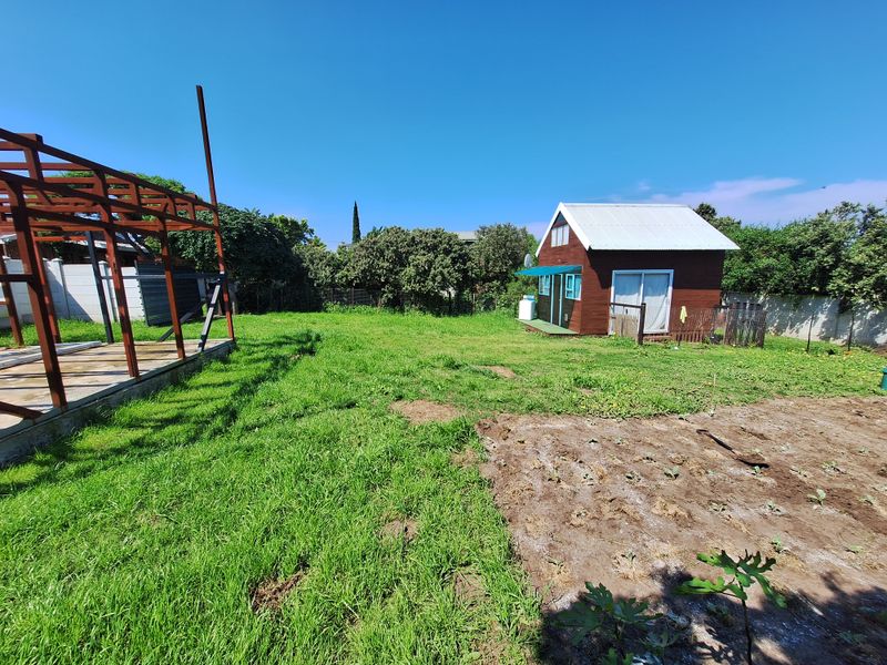 A spacious residential garden featuring a small wooden cabin, an unfinished timber frame structure, and a mix of lawn and cultivated soil areas.