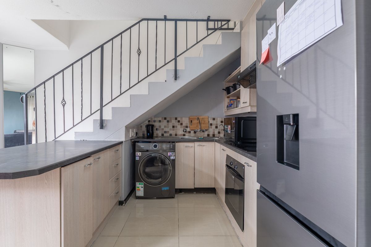 Compact kitchen featuring light wood cabinetry, dark laminate countertops, tiled backsplash, and integrated appliances, efficiently utilizing space beneath the staircase.