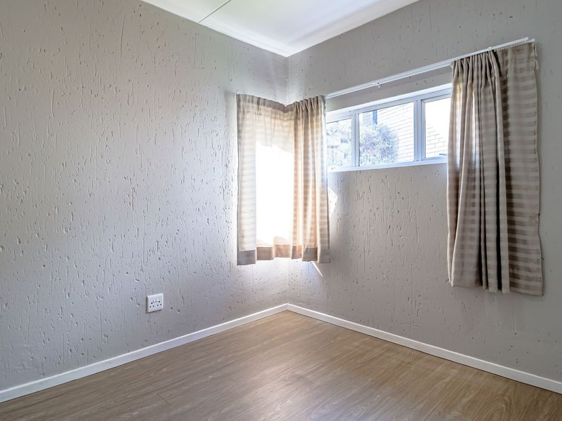 A bright, empty bedroom featuring light-toned laminate flooring, textured walls, and a corner window with neutral curtains.