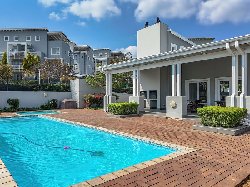 A well-maintained outdoor pool area featuring a paved terrace, covered patio with a built-in braai, and landscaped surroundings within an apartment complex.
