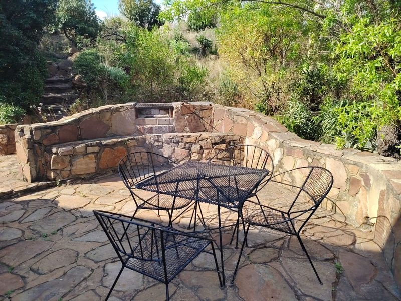 Outdoor patio area featuring natural stone paving, a built-in stone braai, and a stone retaining wall set against a natural garden backdrop.