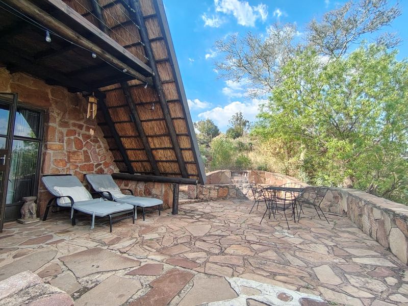 A rustic outdoor patio featuring stone paving, stone wall cladding, and a thatched roof overhang, complete with a built-in stone braai area.