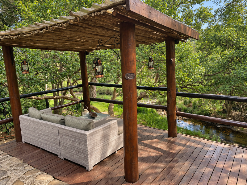 A rustic timber-decked patio featuring a pergola structure, overlooking a natural stream and lush garden surroundings.