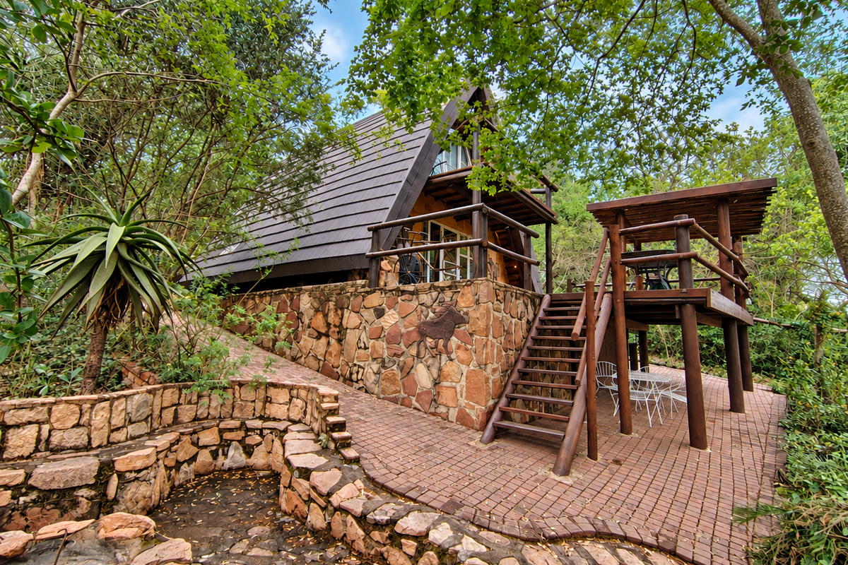 A rustic A-frame townhouse featuring stone cladding, a raised timber deck, and a paved patio area set within a natural, wooded garden environment.