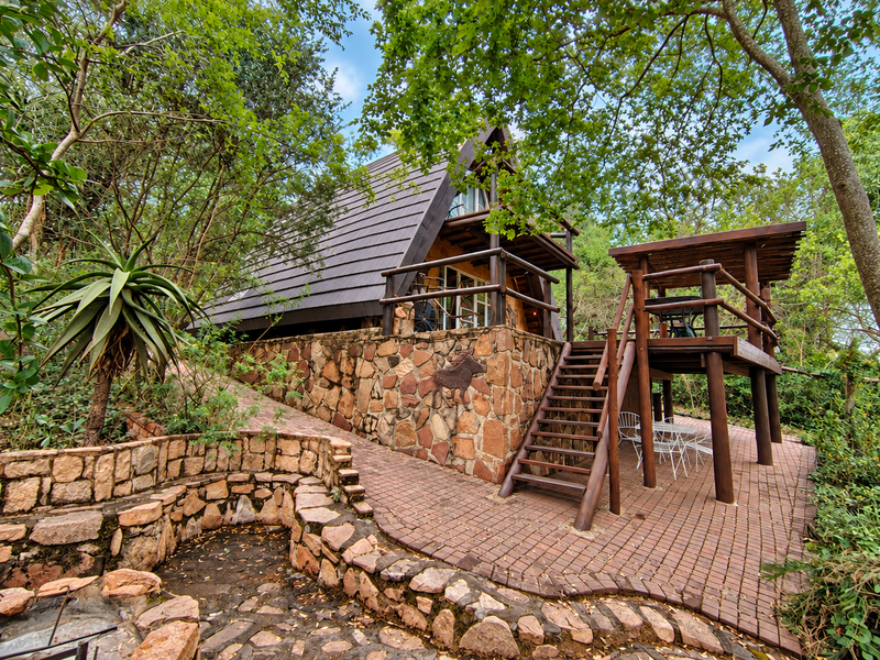 A rustic A-frame townhouse featuring stone cladding, a raised timber deck, and a paved patio area set within a natural, wooded garden environment.