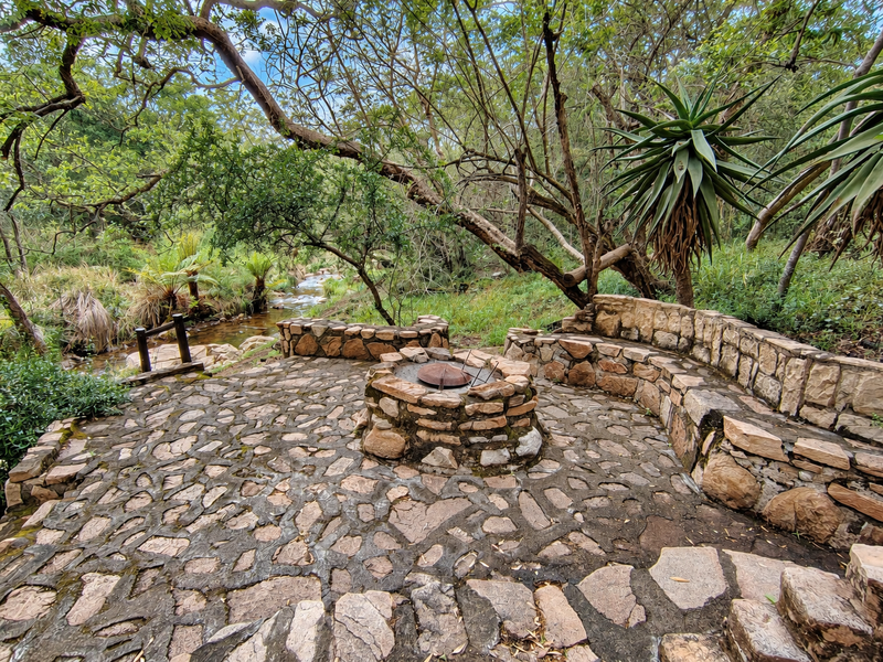 A rustic outdoor patio area featuring a built-in stone fire pit and curved stone bench seating, set in a natural, wooded riverside environment.