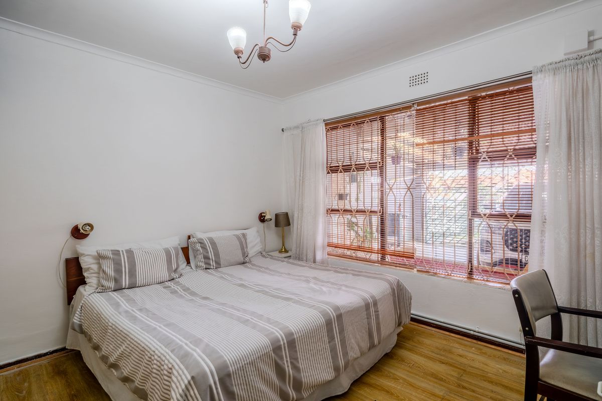 A well-lit bedroom featuring laminate flooring, decorative ceiling coving, and a large window fitted with security gates and blinds.