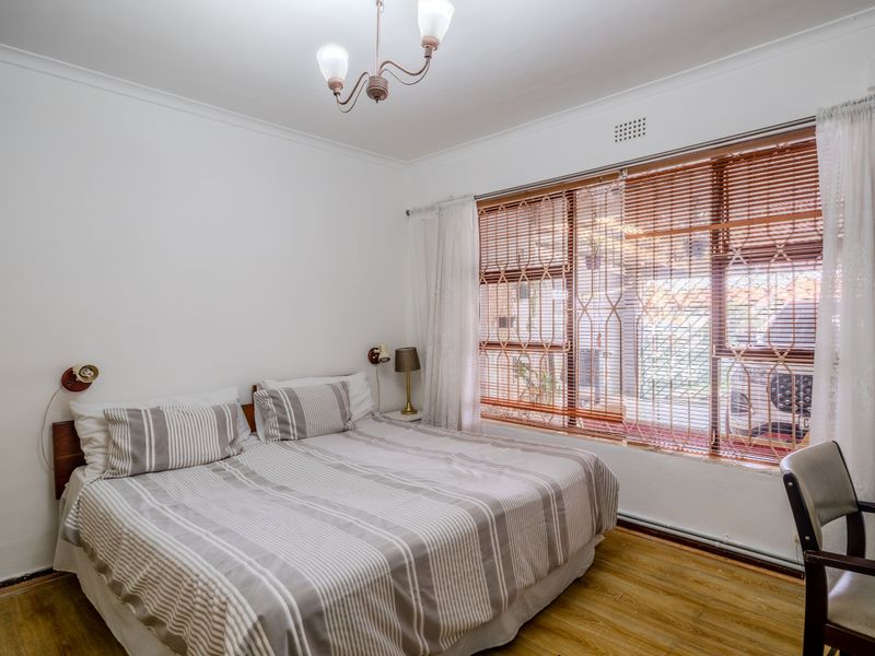 A well-lit bedroom featuring laminate flooring, decorative ceiling coving, and a large window fitted with security gates and blinds.