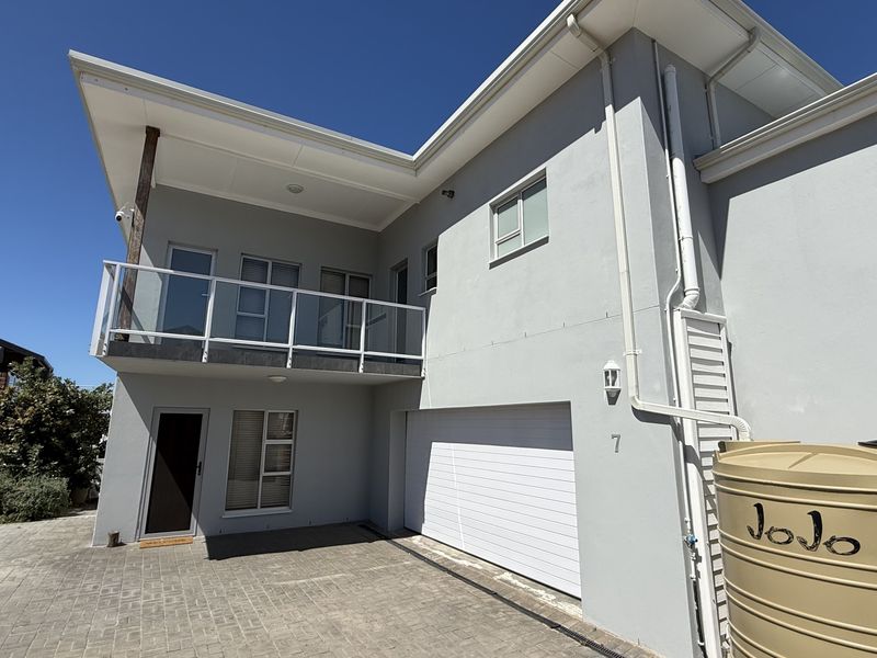Front elevation of a modern two-story house featuring a paved driveway, integrated garage, upper-level balcony with glass balustrade, and a water storage tank.
