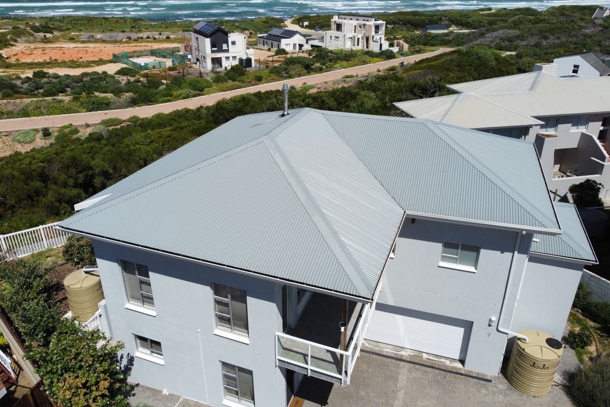 Aerial view of a modern two-story house featuring a corrugated metal roof, paved driveway, garage, and exterior water storage tanks near the coast.