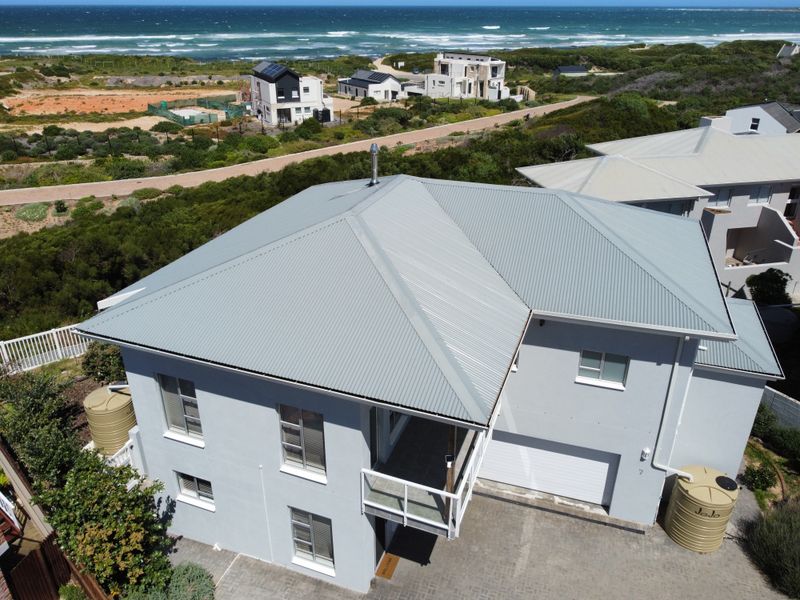 Aerial view of a modern two-story house featuring a corrugated metal roof, paved driveway, garage, and exterior water storage tanks near the coast.
