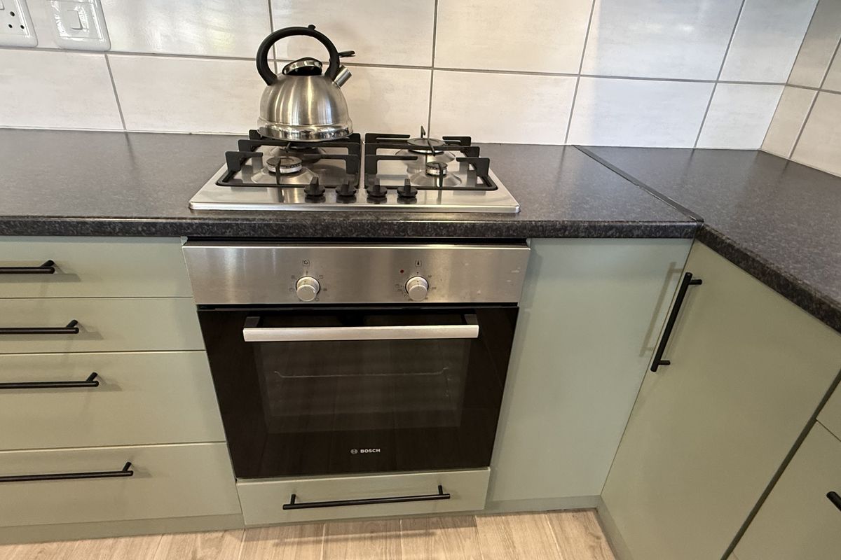 A modern kitchen section featuring a built-in gas hob, stainless steel oven, dark laminate countertops, and sage green cabinetry with matte black hardware.