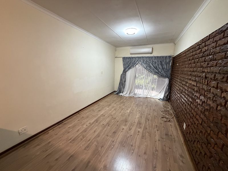 An empty living room featuring laminate flooring, a prominent exposed brick wall, and a wall-mounted air conditioning unit with access to an outdoor area.