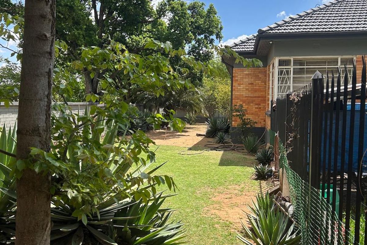 A view of the rear garden featuring a brick exterior, tiled roof, and established landscaping with metal security fencing along the property boundary.