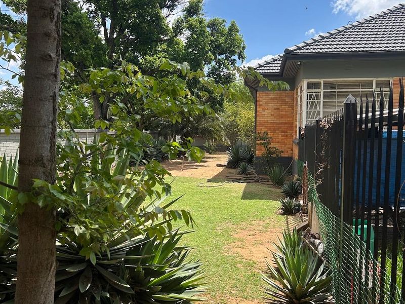 A view of the rear garden featuring a brick exterior, tiled roof, and established landscaping with metal security fencing along the property boundary.