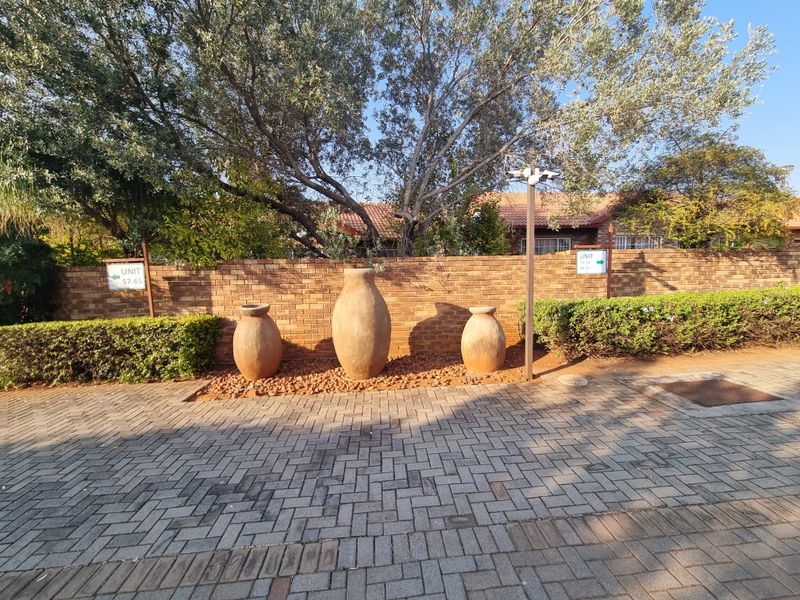 A well-maintained exterior entrance area featuring a brick boundary wall, paved driveway, and established landscaping within the townhouse complex.