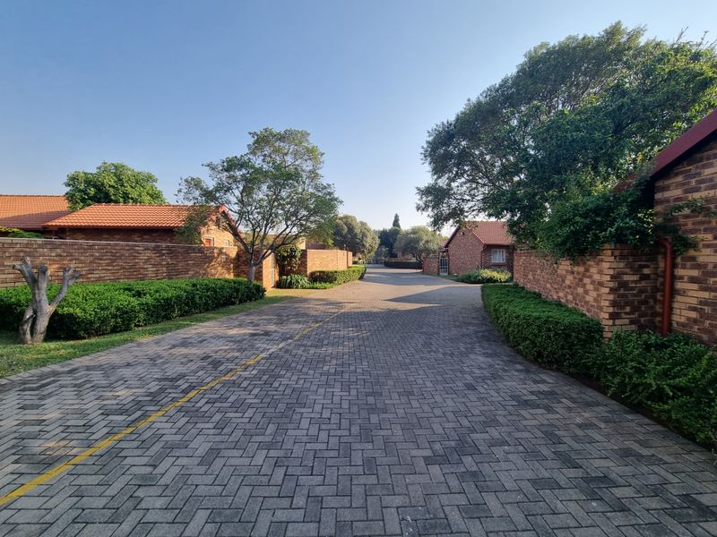 A view of the complex driveway featuring brick paving, established hedges, and surrounding townhouse units with traditional tiled roofs.