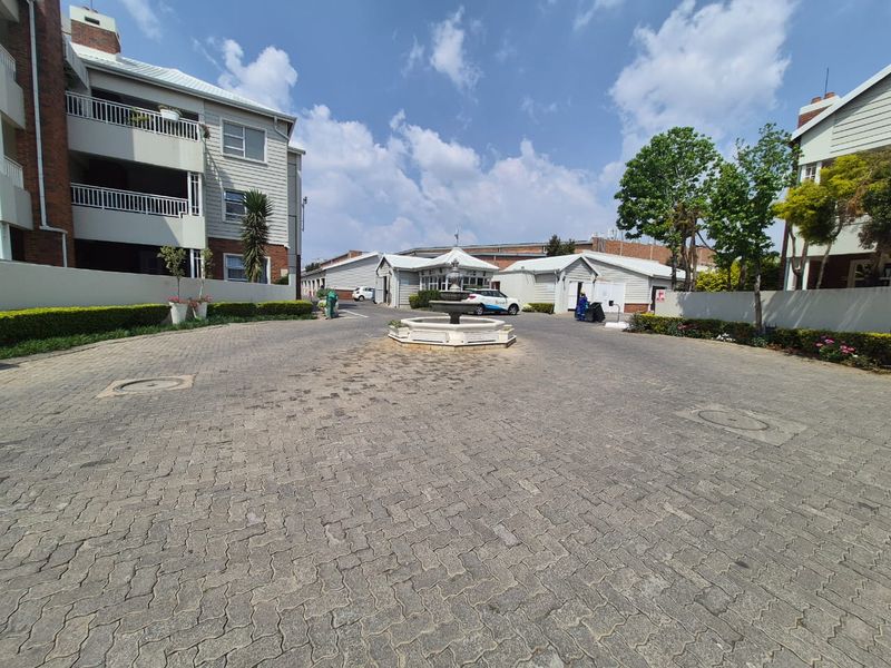 A wide-angle view of the apartment complex entrance featuring a paved driveway, central decorative fountain, and landscaped garden borders.