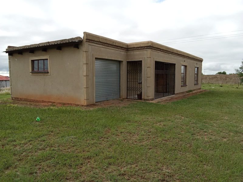 Single-story residential house featuring a roller garage door, security gates, and plastered exterior walls, situated on a large open plot.