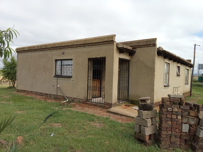 A single-story residential property featuring plastered exterior walls, security gates, and metal window frames, currently requiring significant maintenance and exterior finishing.