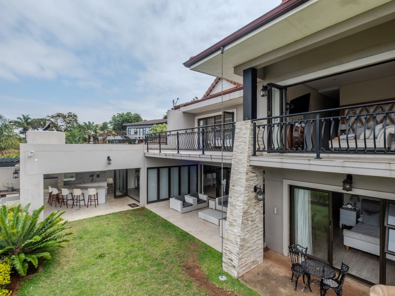 Spacious rear exterior featuring a covered patio with built-in kitchen, upper balcony with wrought iron railings, and landscaped garden access.