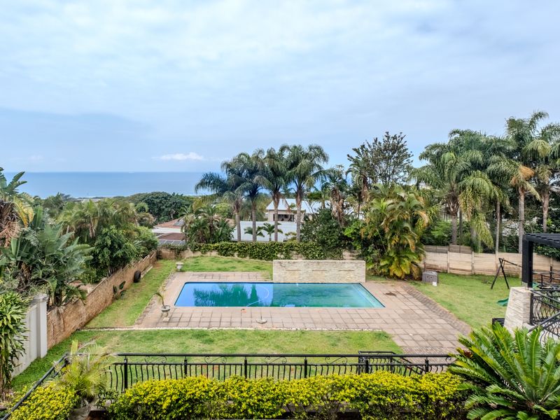 Elevated view of a backyard featuring a swimming pool with a stone water feature, paved surround, and distant ocean views.