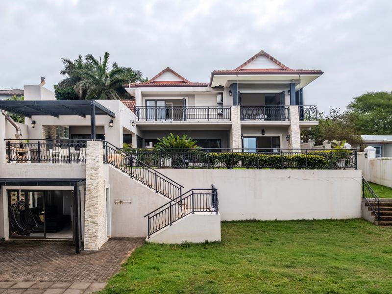 Rear view of a multi-level house featuring a spacious balcony, paved patio area with built-in braai, and well-maintained garden space.