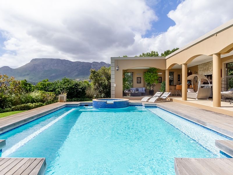 A well-maintained swimming pool with a raised spa, timber decking, and a spacious covered patio area featuring arched columns and mountain views.