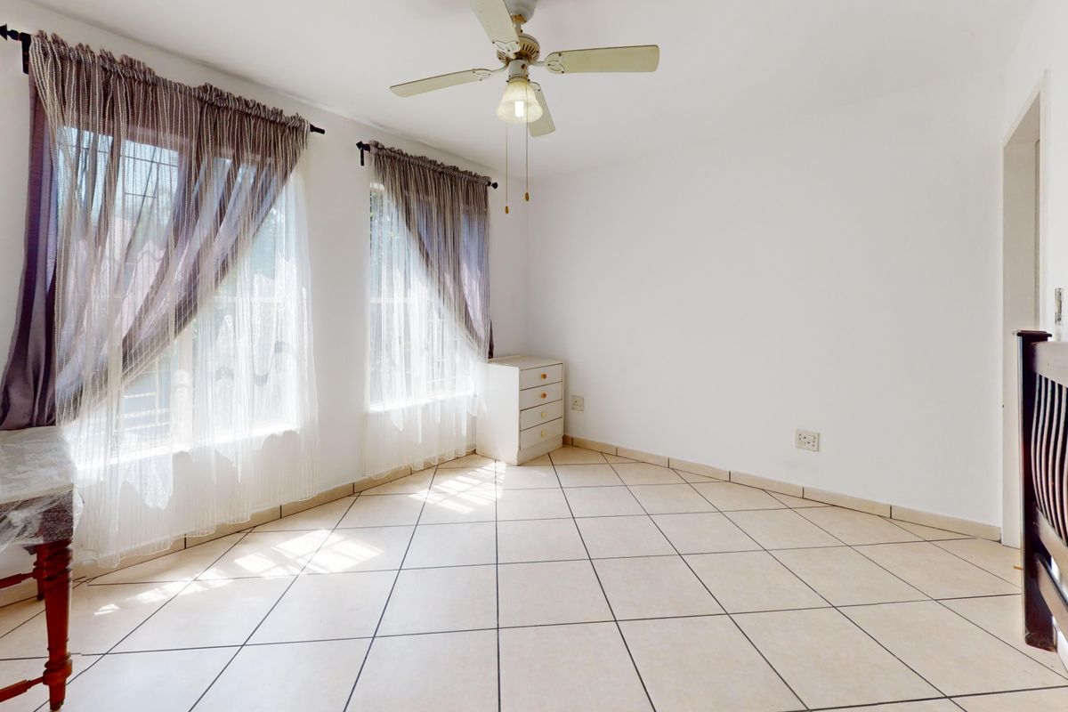 A bright, empty bedroom featuring neutral tiled flooring, a ceiling fan, and large windows with security bars.