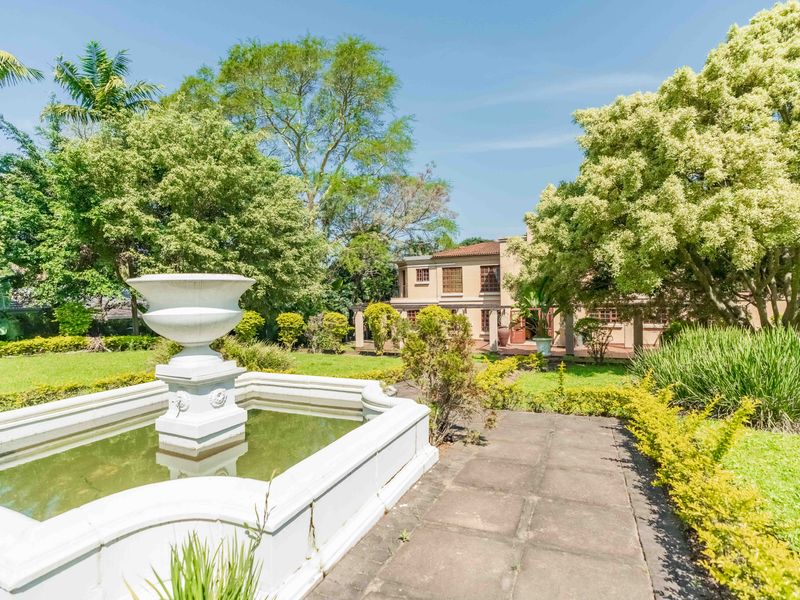 A well-maintained garden featuring a central stone fountain, paved pathways, and mature landscaping leading to the rear facade of the house.