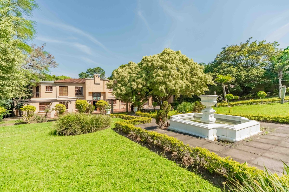 A spacious rear garden featuring a decorative stone fountain, paved pathways, and mature landscaping set against the rear elevation of the two-story residence.