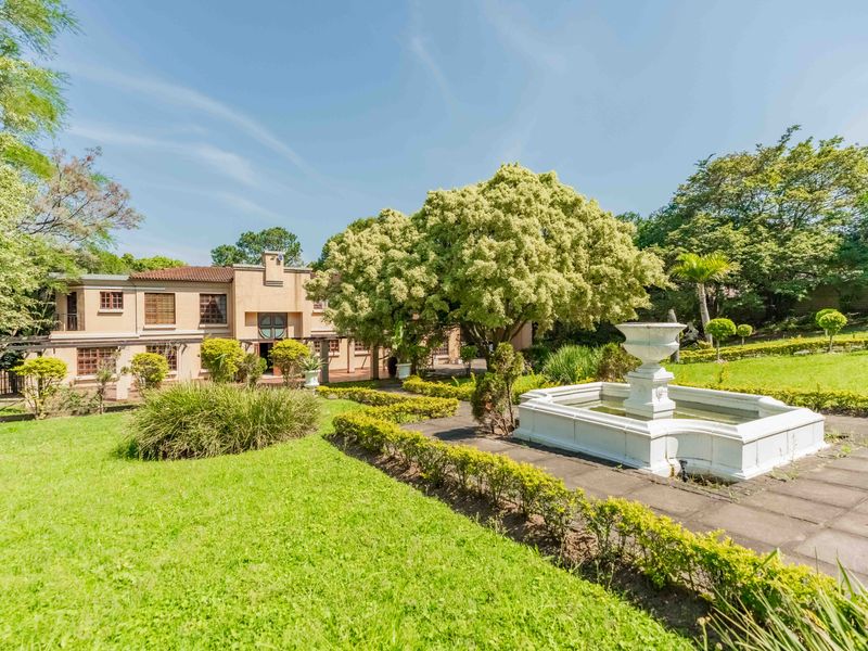 A spacious rear garden featuring a decorative stone fountain, paved pathways, and mature landscaping set against the rear elevation of the two-story residence.
