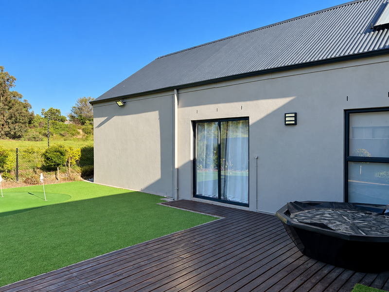 Rear exterior view featuring a modern home with a corrugated metal roof, timber decking, artificial lawn, and integrated exterior lighting.