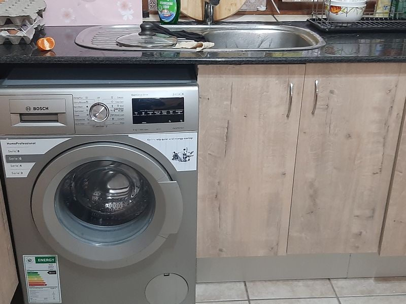 A functional kitchen area featuring light wood-finish cabinetry, dark granite countertops, a stainless steel sink, and a built-in washing machine.