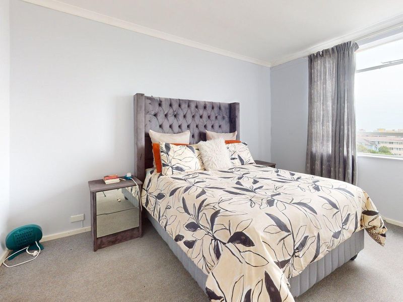 A well-lit bedroom featuring neutral wall tones, carpeted flooring, and a large window providing natural light and an urban view.
